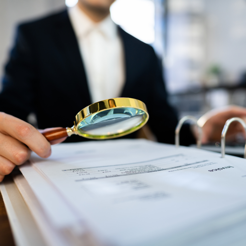 Person examining documents with a magnifying glass, representing detailed analysis for a marketing audit report to improve online business performance.